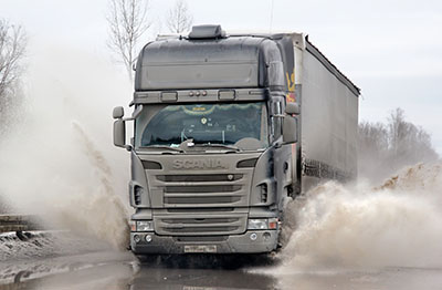 A large truck driving through heavy standing water on the road, splashing waves of water up on either side of it.