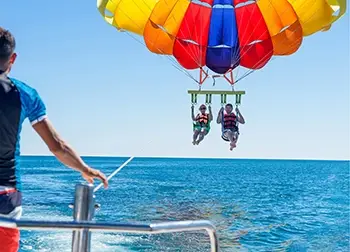 A couple fly in a tandem parasail with a colorful canopy wing, towed by a boat, while their operator watches from the boat deck.