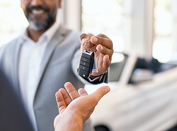 A car dealer hands over the keys to a new car to its owner, with the car in the background.